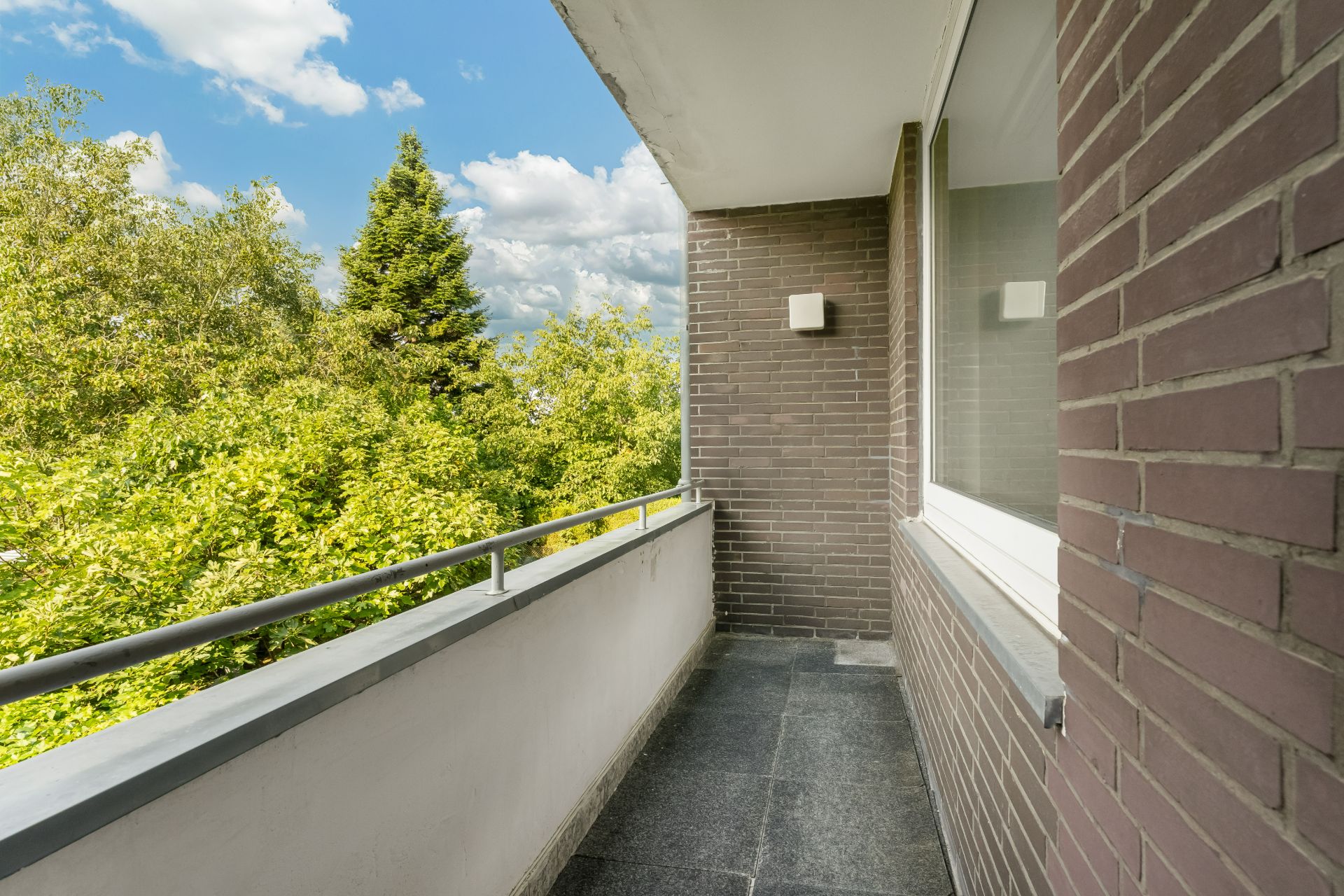 Schmaler Balkon mit Fliesenboden und Blick auf grüne Bäume, angrenzend an eine Backsteinwand.