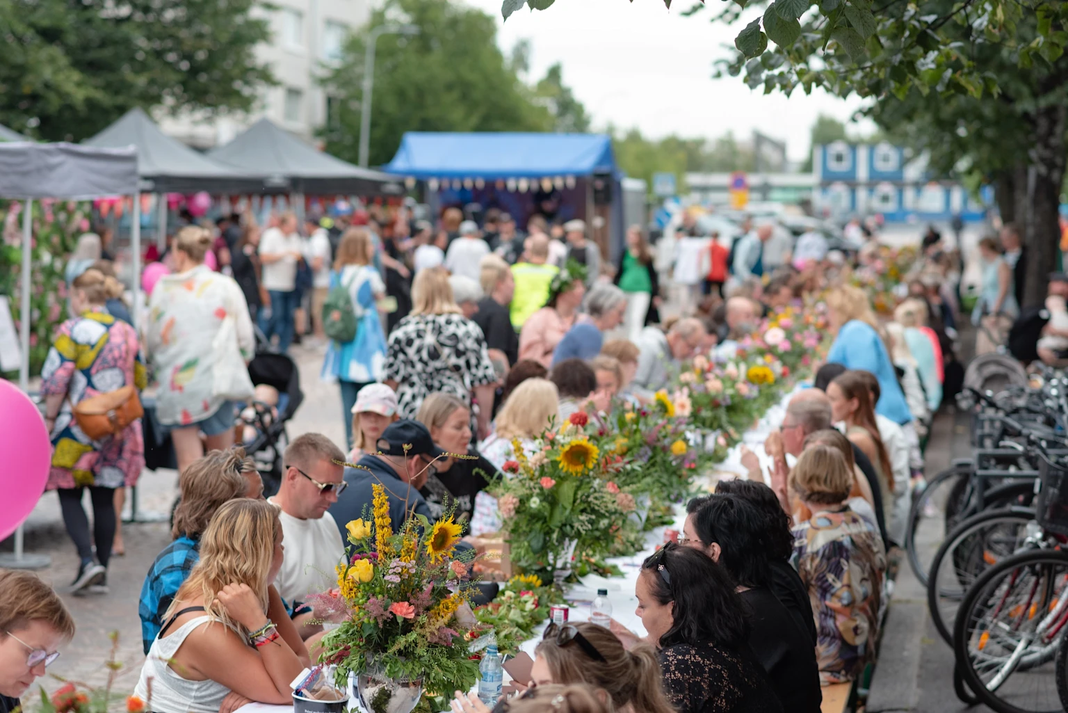 Kesäisesti pukeutuneita ihmisiä istumassa pitkän ruokapöydän ääressä Oulun keskustan kadulla. Ruokapöytä on koristeltu kesäkukilla. Taustalla näkyy tapahtuman ruokakojuja. Kuva: Kevin Kallombo, Oulu2026