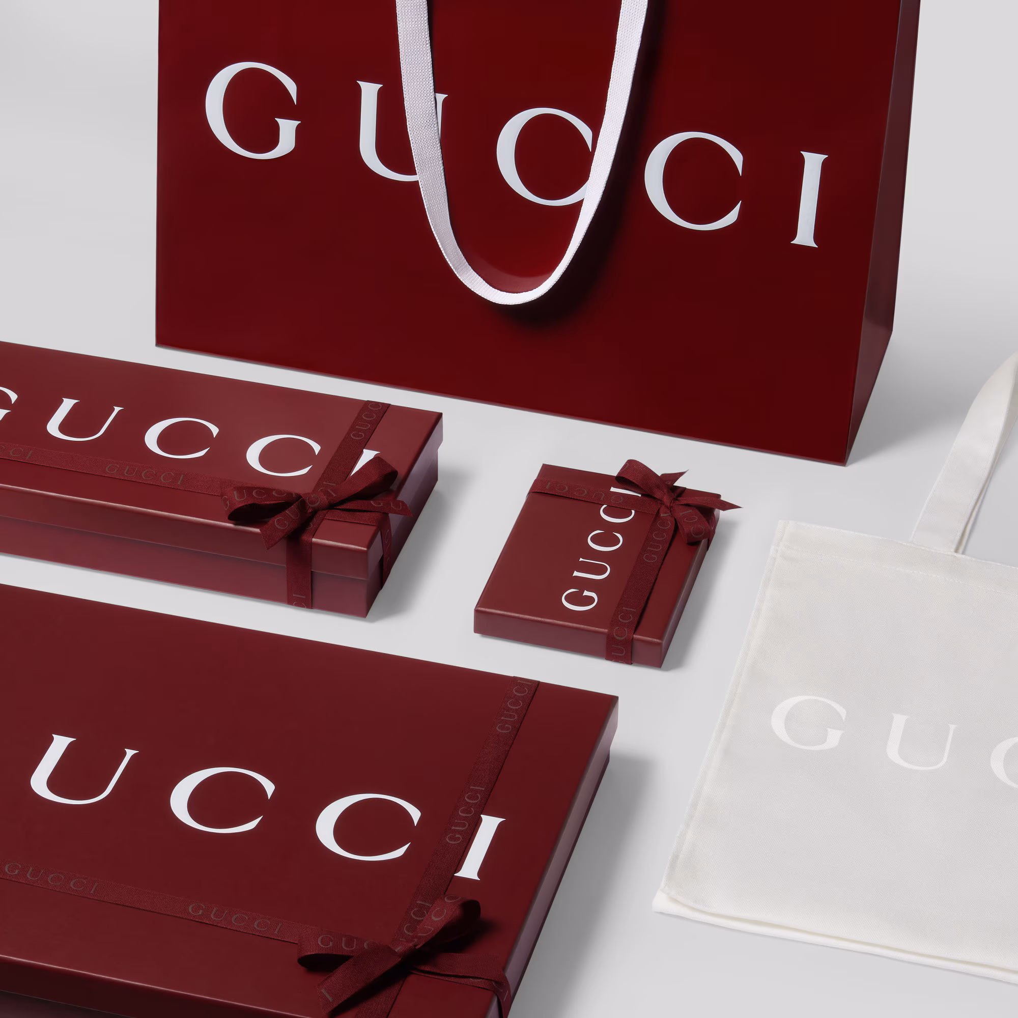 Gucci red shopping bag surrounded by matching different sizes of Gucci signature packaging and a white cotton canvas bag with Gucci logo in white.