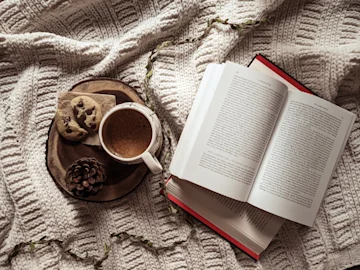A mug of hot chocolate, broken cookie and pine cone on a plate next to an open book on a blanket. 