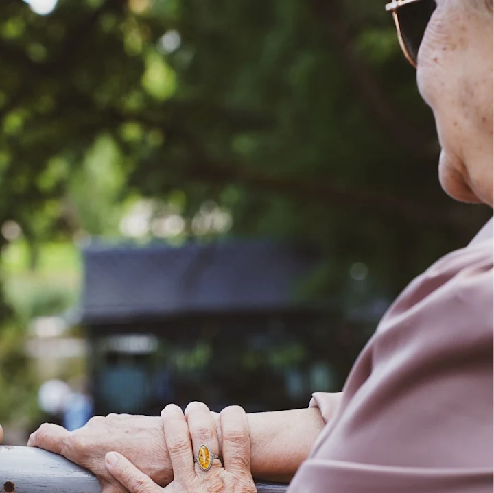 An older woman with glasses looking out over a garden on a sunny day.