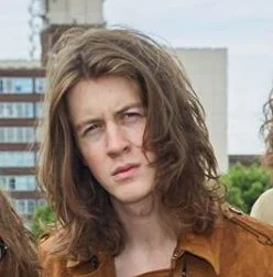 Tom Ogden from the band Blossoms, with long brown hair and wearing a brown leather-looking jacket, looking directly at the camera.