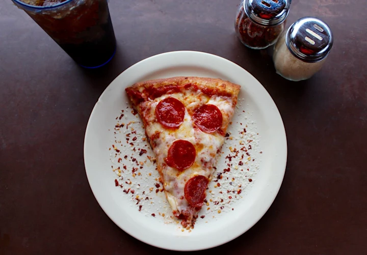 A slice of pepperoni pizza on a paper plate. It's on a table below a glass of coke and salt and pepper shakers. 