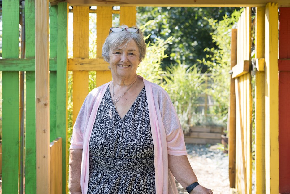 Smiling older woman with grey hair and sunglasses on head, wearing pink cardigan, standing in colourful wooden structure.