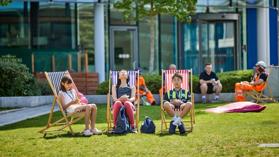 Three teenagers sit on deck chairs on a patch of grass outside a glass office building at Stockport Exchange. Behind them is is a workman in a florescent orange outfit and hard hat, talking to three others sitting in front of a hedge. It is a bright, sunny day.