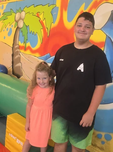 A teenager and Autisk youth volunteer poses with a young girl in a room with bright illustrations on the wall. They're both smiling.