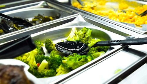 Serving spoon with broccoli, amongst trays of other food.