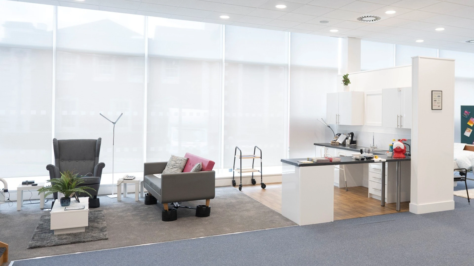 A bright open space with a mock open-plan kitchen and living room. Soft furnishings, tables and kitchen cabinets and countertops are all included, with different assistive equipment on display. 
