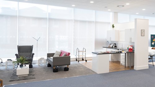 A bright open space with a mock open-plan kitchen and living room. Soft furnishings, tables and kitchen cabinets and countertops are all included, with different assistive equipment on display. 