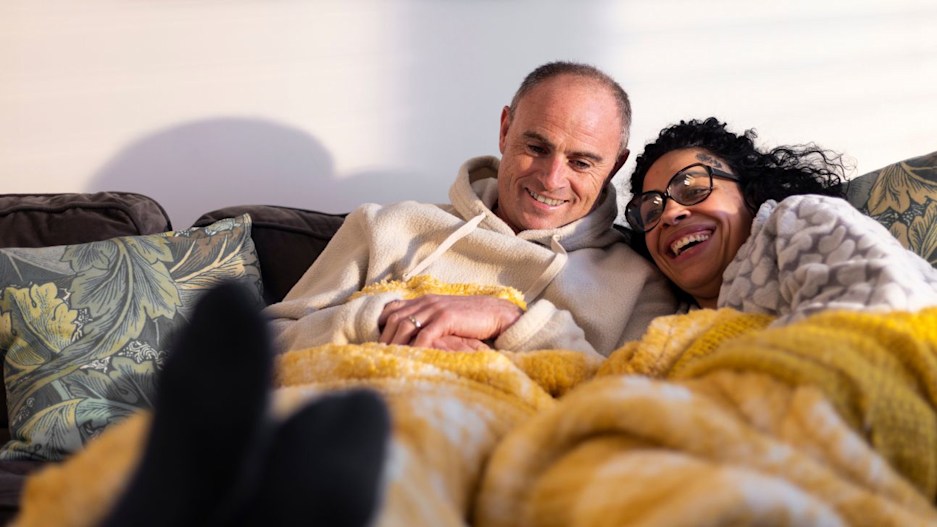 A man and woman sitting under a fleecy blanket, relaxing on a sofa. Their legs are stretched out in front of them and the woman is leaning her head on the man's shoulder. They are both smiling. 