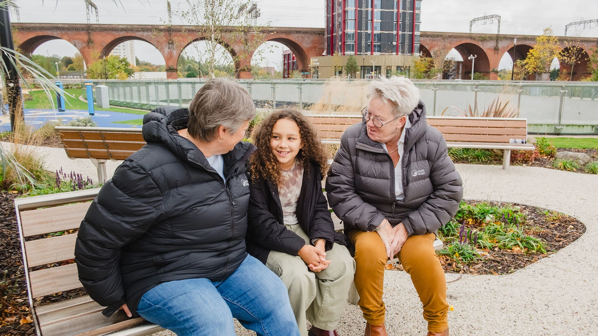 Two women are sat talking with a young girl. They're all smiling, and are sat on a bench with Stockport's viaducts behind them.