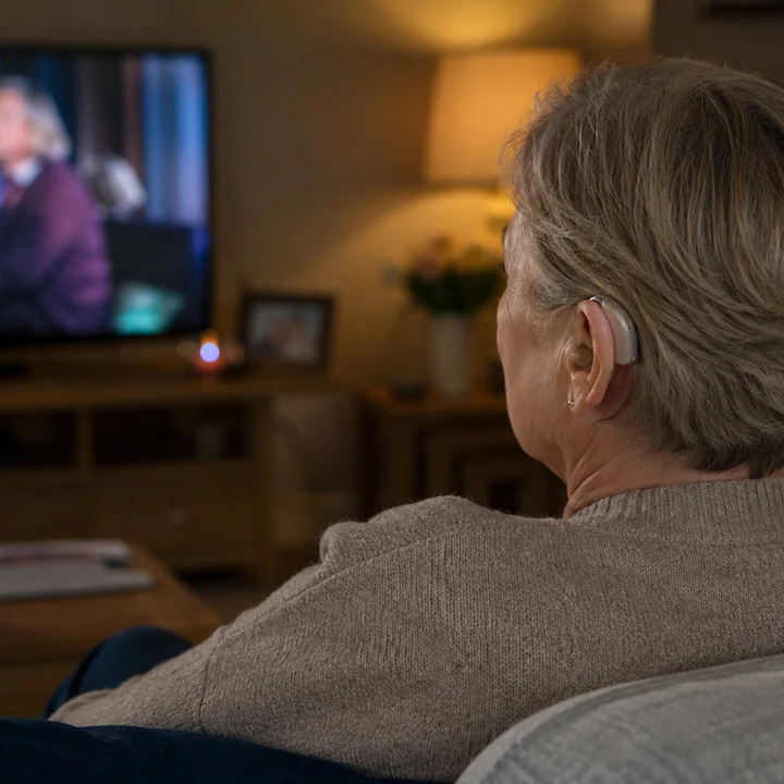 A woman watching television in a comfortable, softly lit home setting. She has short, slightly greying hair and us wearing a cream-coloured jumper. We can only see the back of her head and shoulders, and she's wearing a hearing aid.