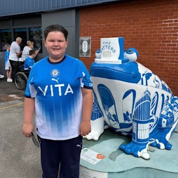 Bertie, a young boy in a Stockport County Football Club shirt, stood smiling by a statue of a frog painted in Stockport County's blue and white colours.