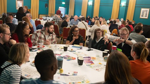 A group of people sat at a round table, engaged in a discussion. One man is talking enthusiastically while everyone else is leaning in to listen. There are sheets of paper and leaflets spread across the table.