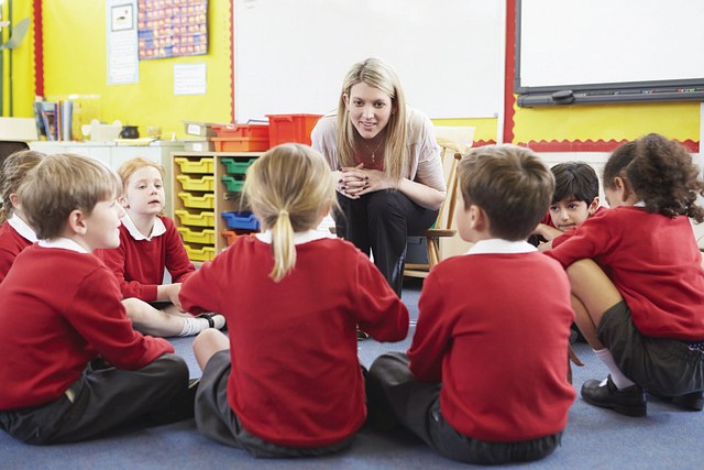 A woman sitting in a bright primary school classroom. She is leaning forward on a chair, smiling and listening to a young girl who is sitting with other pupils in a circle around the woman. 