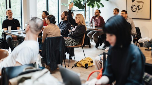 Men and women of different ethnicities sitting together at tables in a meeting room, one person with their hand raised to ask a question.