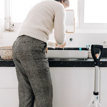 An older woman washing her hands in a sink. There's a piece of assistive equipment next to her.