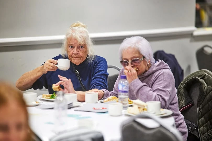Two older women sitting at a table inside with a plate of food in front of them and hot drinks. 