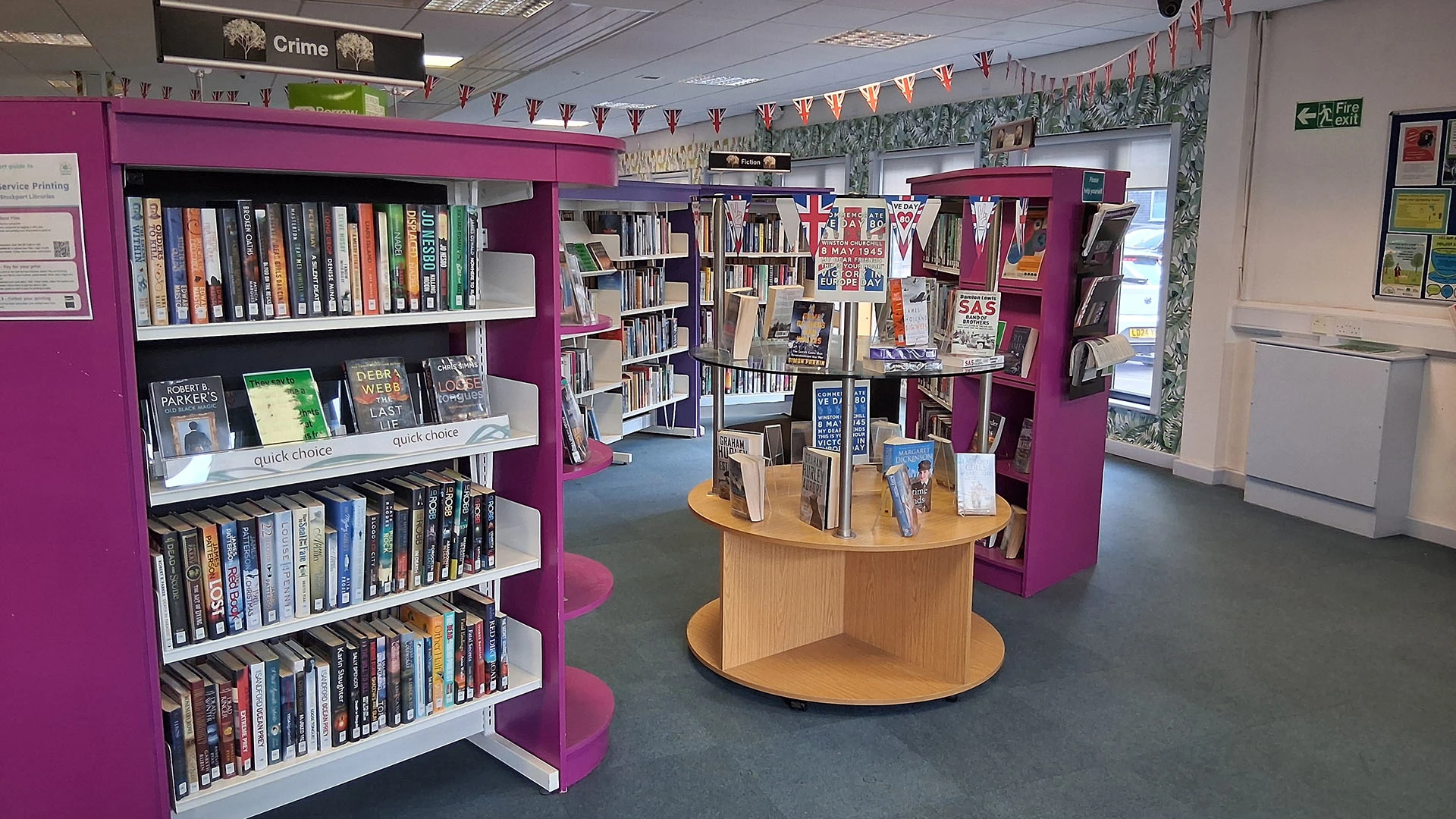 Library interior with purple bookshelves, circular wooden display table, Union Jack bunting, and Crime section signage.