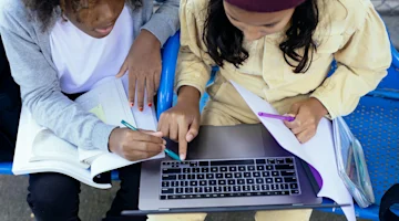 Two people sitting on a bench studying together with a laptop, books, and notebooks.