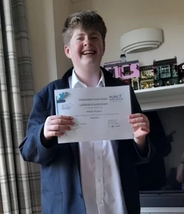 A teenage boy in a living room, smiling wide and posing in front of curtains and a TV,. He is holding a certificate to the camera.