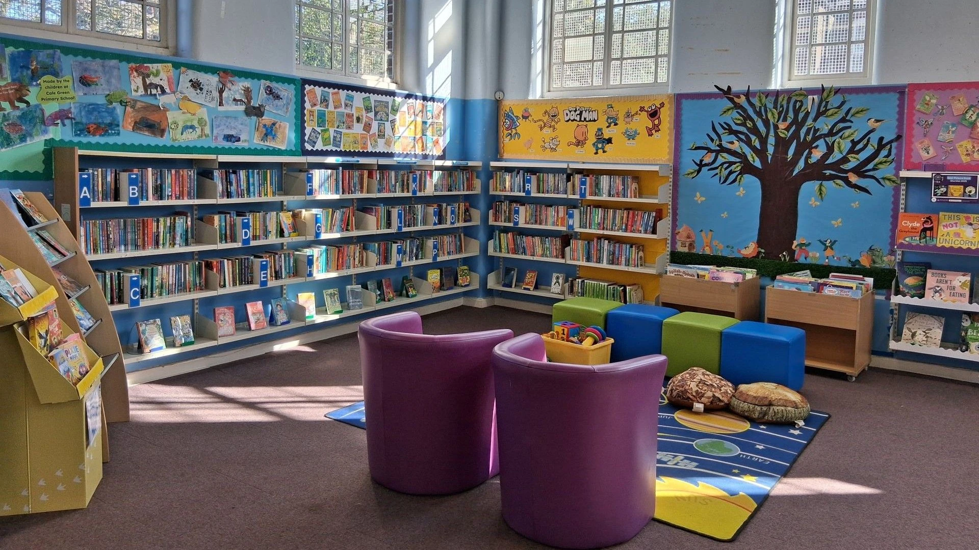 A light, airy room full of colourful furniture, bright drawings on the wall, and shelves full of children's books. A rug is in the centre with furniture arranged around it and the run is shining through the windows.
