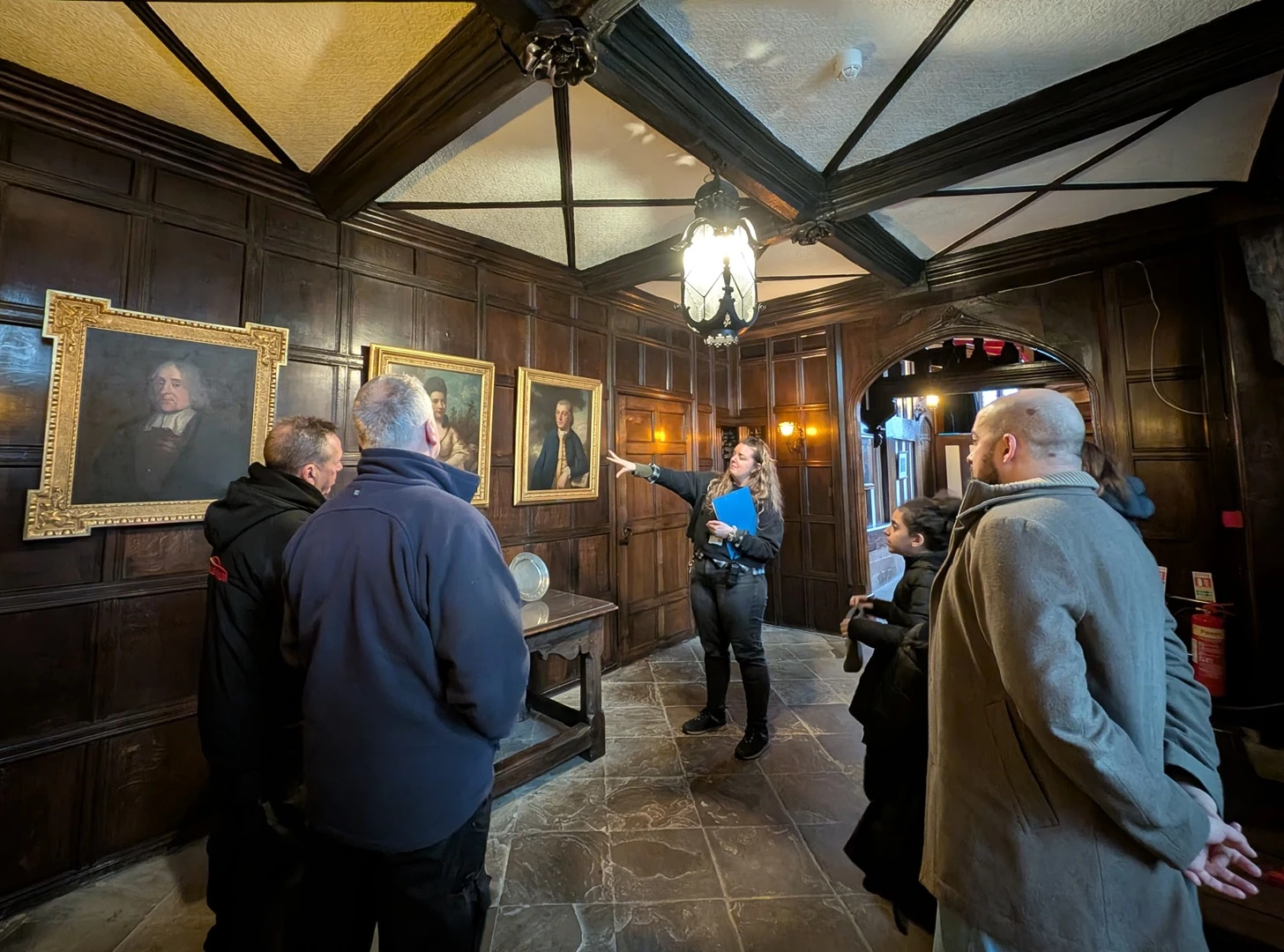 A group stood listening to a tour guide in Bramall Hall. The walls are covered in wooden panels, there are wooden beams on the black and white Tudor-style ceiling, and the room is lit by an ornate hanging lamp. The tour guide, a woman with long blonde hair, is pointing at a row of realistic oil paintings.