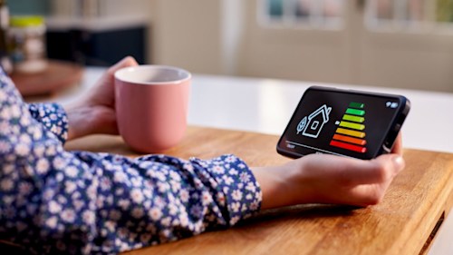 A person's hands holding a smart meter with an energy label and house graphic on the screen in one hand and a mug in the other. Their arms are resting on a table.