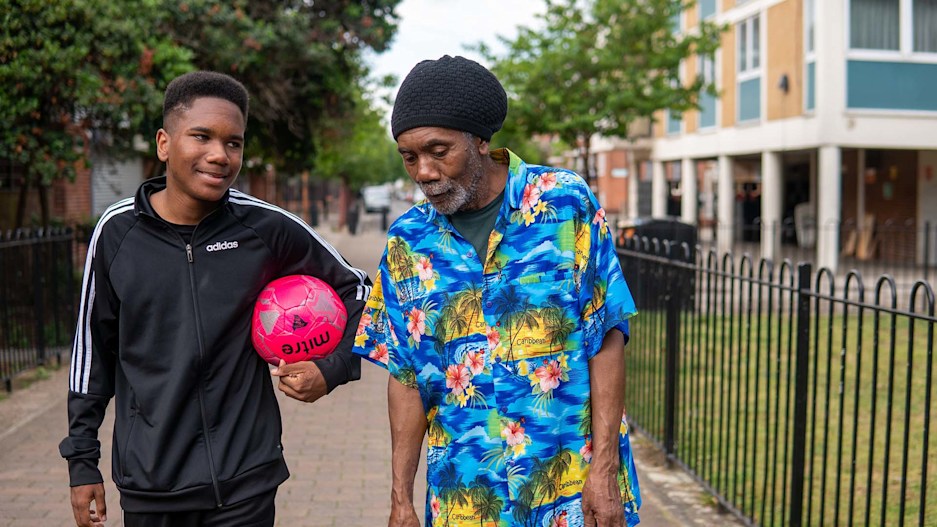 A young man holding a football, walking down the street with an elderly man in a suburban area.