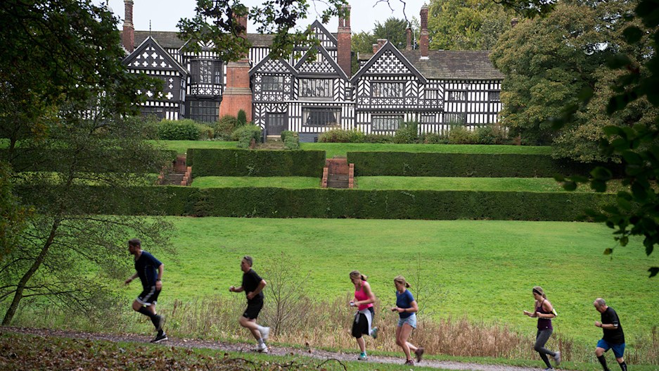 A group of people running past Bramall Hall, an ornate Tudor building in a bright green field.