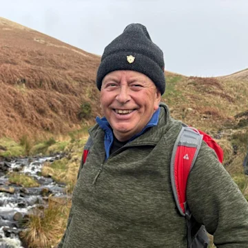 Eric, a man wearing a woolly hat, fleece and backpack, smiling at the camera. He's stood in an outdoor setting, with hills and a stream behind him.