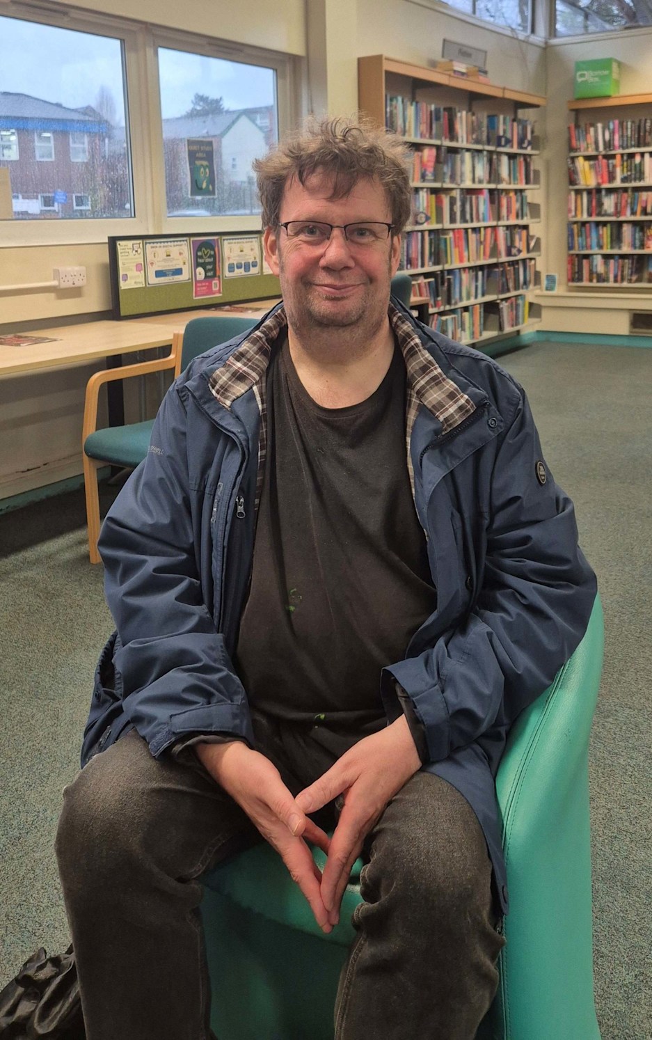 Stephen sitting in an armchair in Hazel Grove Library, a large room with two bookcases full of books behind him and a desk beneath two windows. 