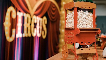A popcorn machine in the foreground with a large circus sign and red curtains in the background.
