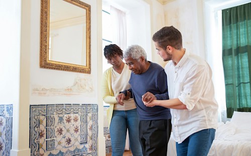 A man and a woman holding the hands and arms of an older man, helping him to walk.
