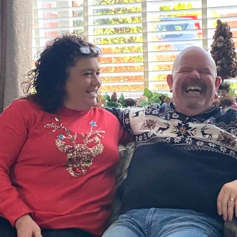 A man and a woman (Les and Debbie) sat on a sofa wearing Christmas jumpers. They are smiling and laughing.