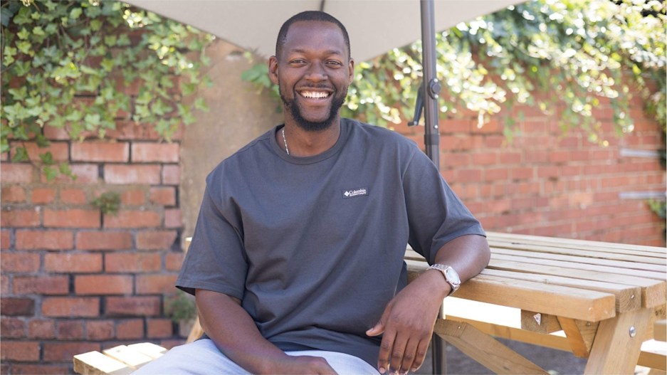 Man in dark t-shirt sitting casually at a wooden picnic bench on a bright, sunny day. He's smiling at the camera.