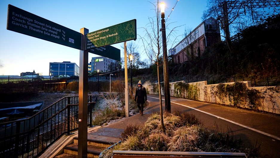 A woman in winter clothing including a woolly hat walking towards the steps down to the town centre along the link road from Viaduct Park to Stockport Rail Station. A pedestrian road sign is in the foreground, and frost can be seen on the sign and plants. The sky is a dusky blue, as though early morning or early evening.