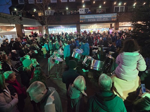 A crowd gathers around a band playing steel drums in front of a large Christmas tree.