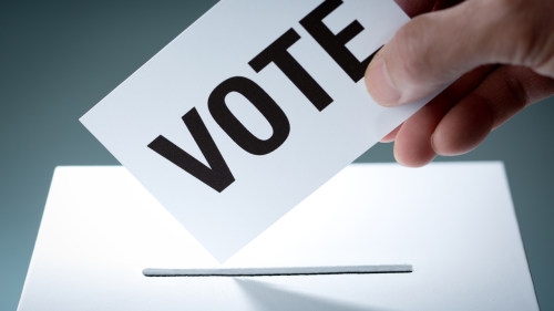 A hand places a card in a ballot box. On the card is written the word 'VOTE' in large block capitals.