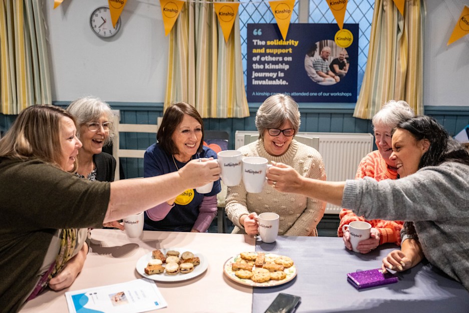 Six women sitting around a table, reaching across to clink mugs of tea or coffee together. They're all smiling. On the table are plates of biscuits. A kinship carer poster and bunting is on the wall behind them.  