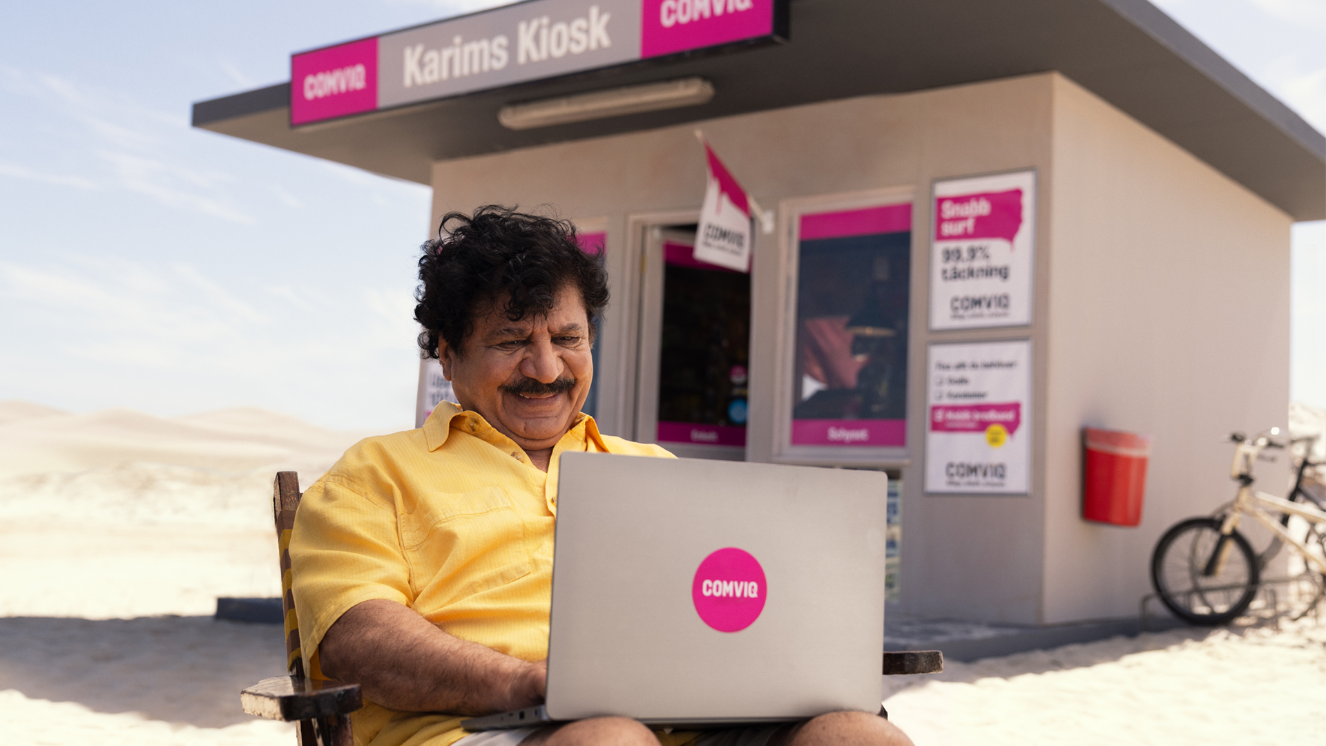 Karim sitter med sin laptop i knät utanför Comviq-kiosken och ser nöjd ut