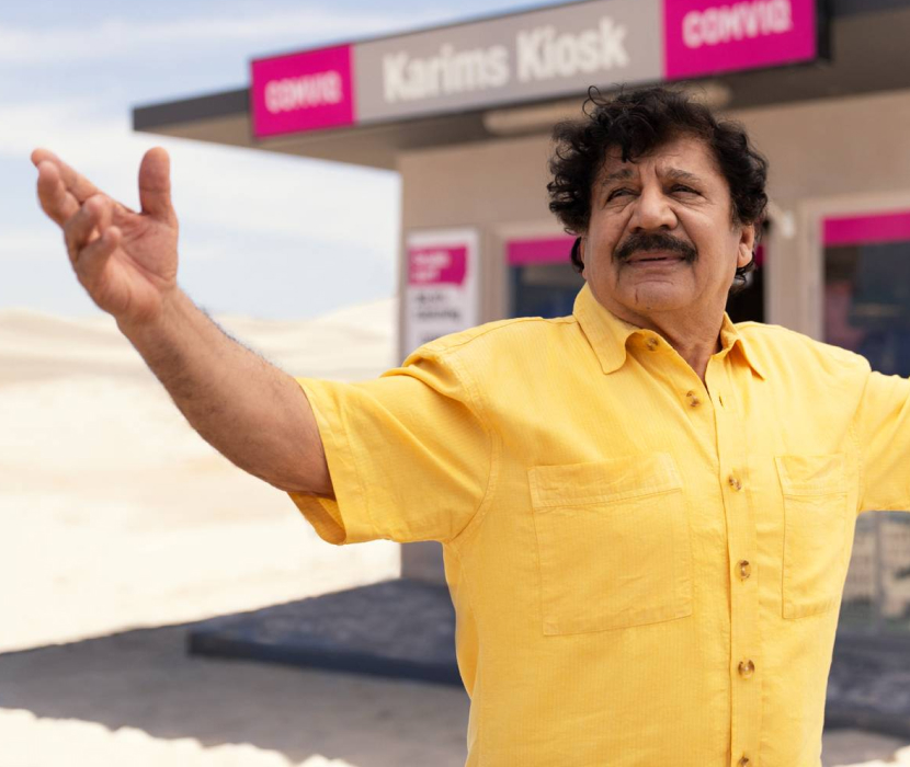 Person i gul skjorta gestikulerar framför Karims Kiosk med rosa Comviq-skyltar i bakgrunden.