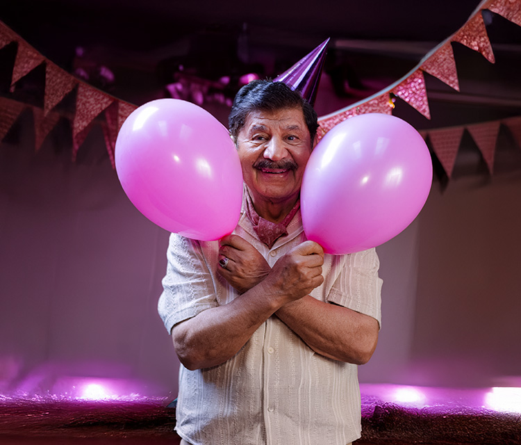 En leende äldre man med partyhatt håller två rosa ballonger omgivna av rosa vimplar i bakgrunden