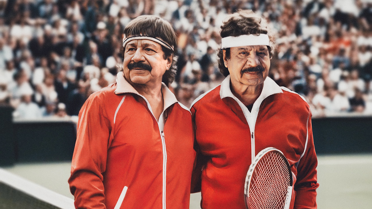 Två äldre män med mustascher i röda träningsjackor och pannband står på en tennisbana med publik i bakgrunden.