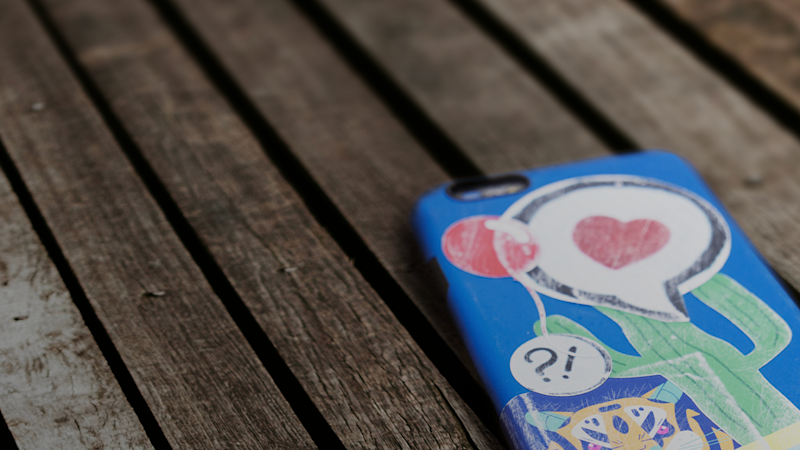 Gammal mobiltelefon med baksidan uppåt på träbord, slitet mobilskal med kaktus, hjärta och andra figurer.