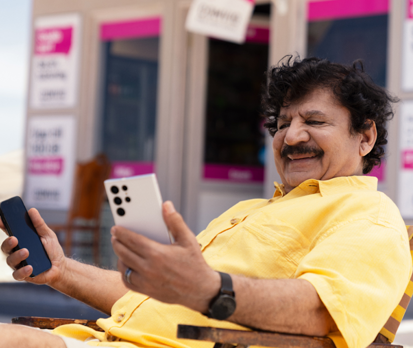 Karim sitter tillbakalutad i en stol utanför kiosken och tittar på en av två mobiler som han håller i vardera handen