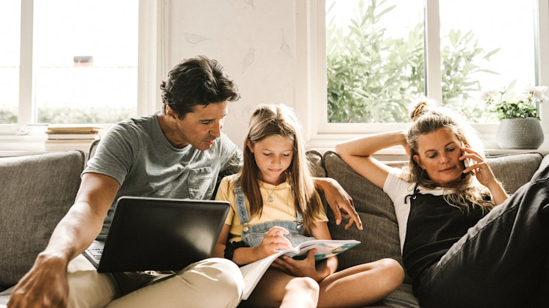 Familj i soffa framför fönster, pappa med laptop, flicka läser tidning och kvinna pratar i telefon. Ljust rum med gröna växter utanför.