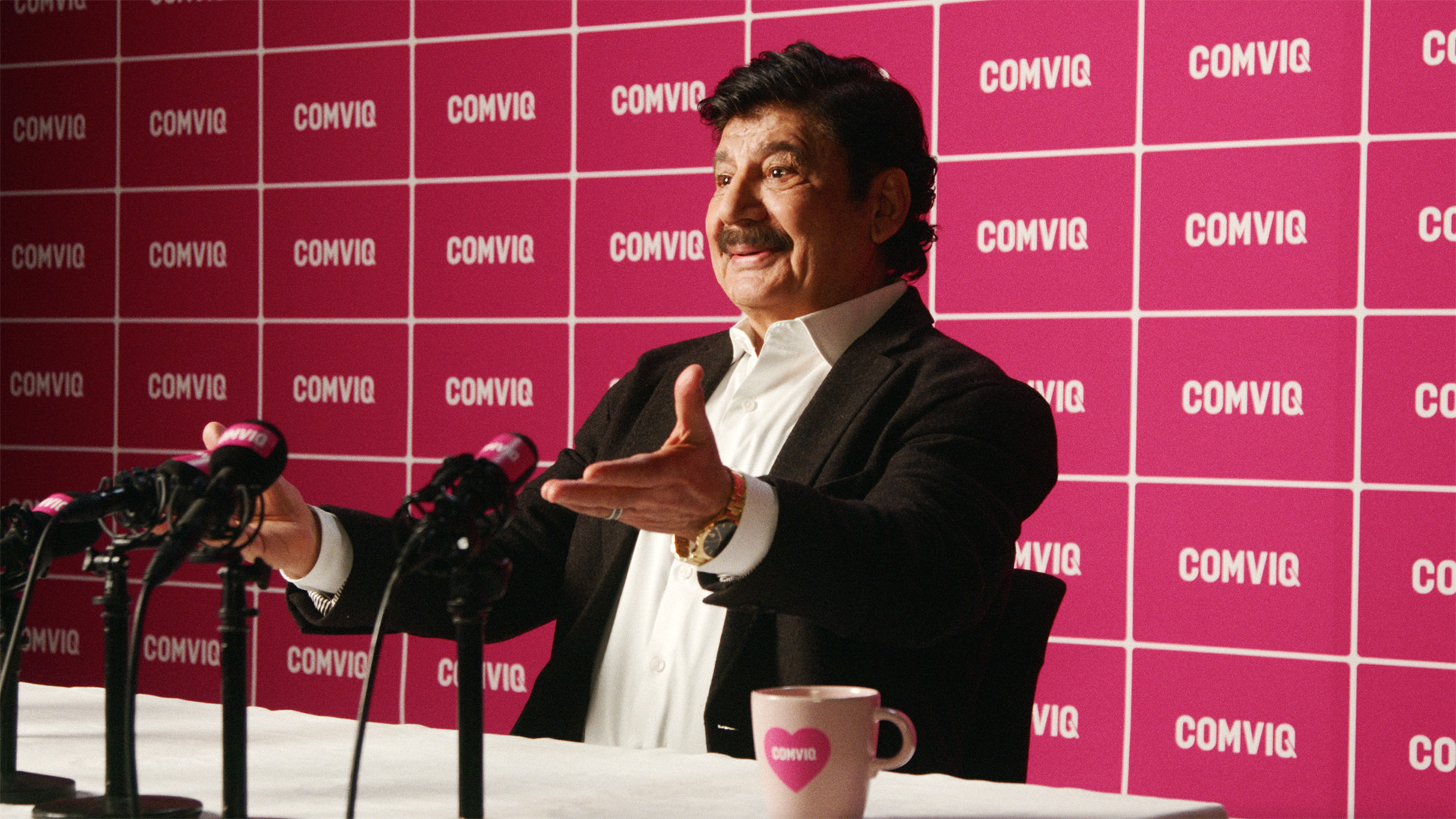 Karim i kostym talar vid presskonferens framför rosa COMVIQ-bakgrund med mikrofoner i förgrunden.