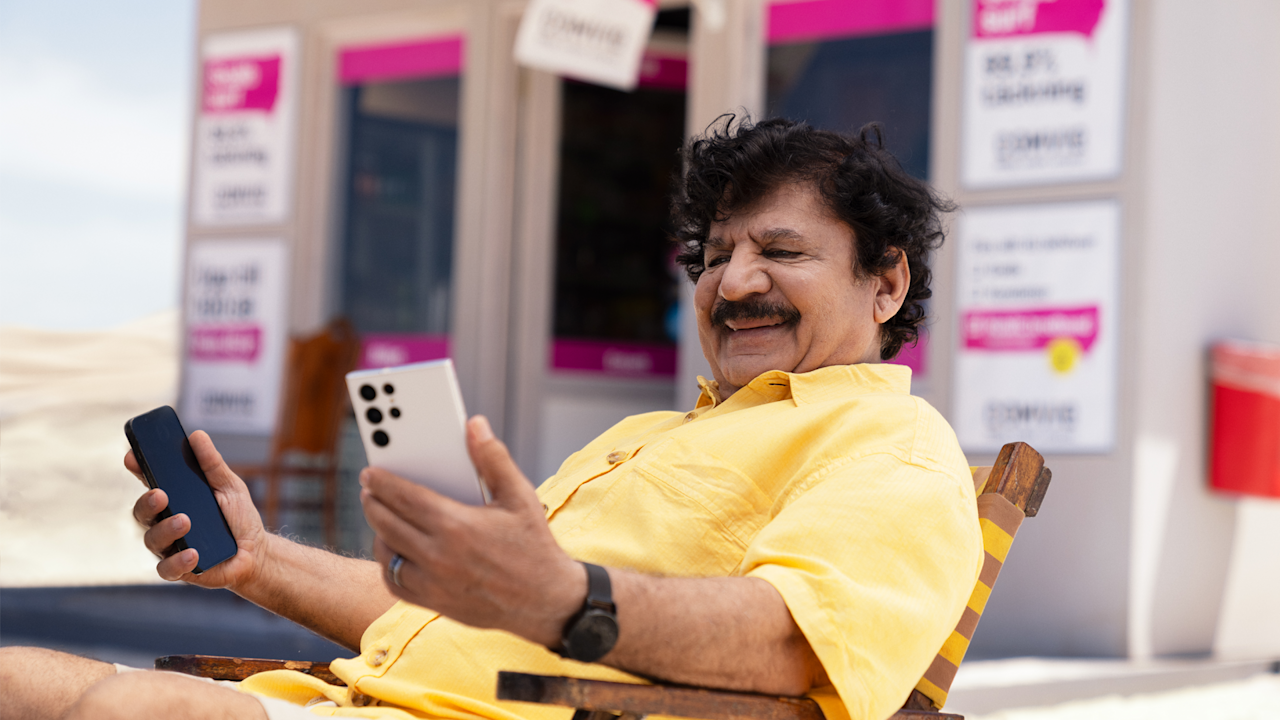 Karim sitter tillbakalutad i en stol utanför kiosken och tittar på en av två mobiler som han håller i vardera handen