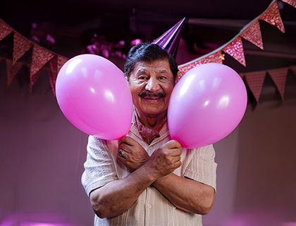 En leende äldre man med partyhatt håller två rosa ballonger omgivna av rosa vimplar i bakgrunden